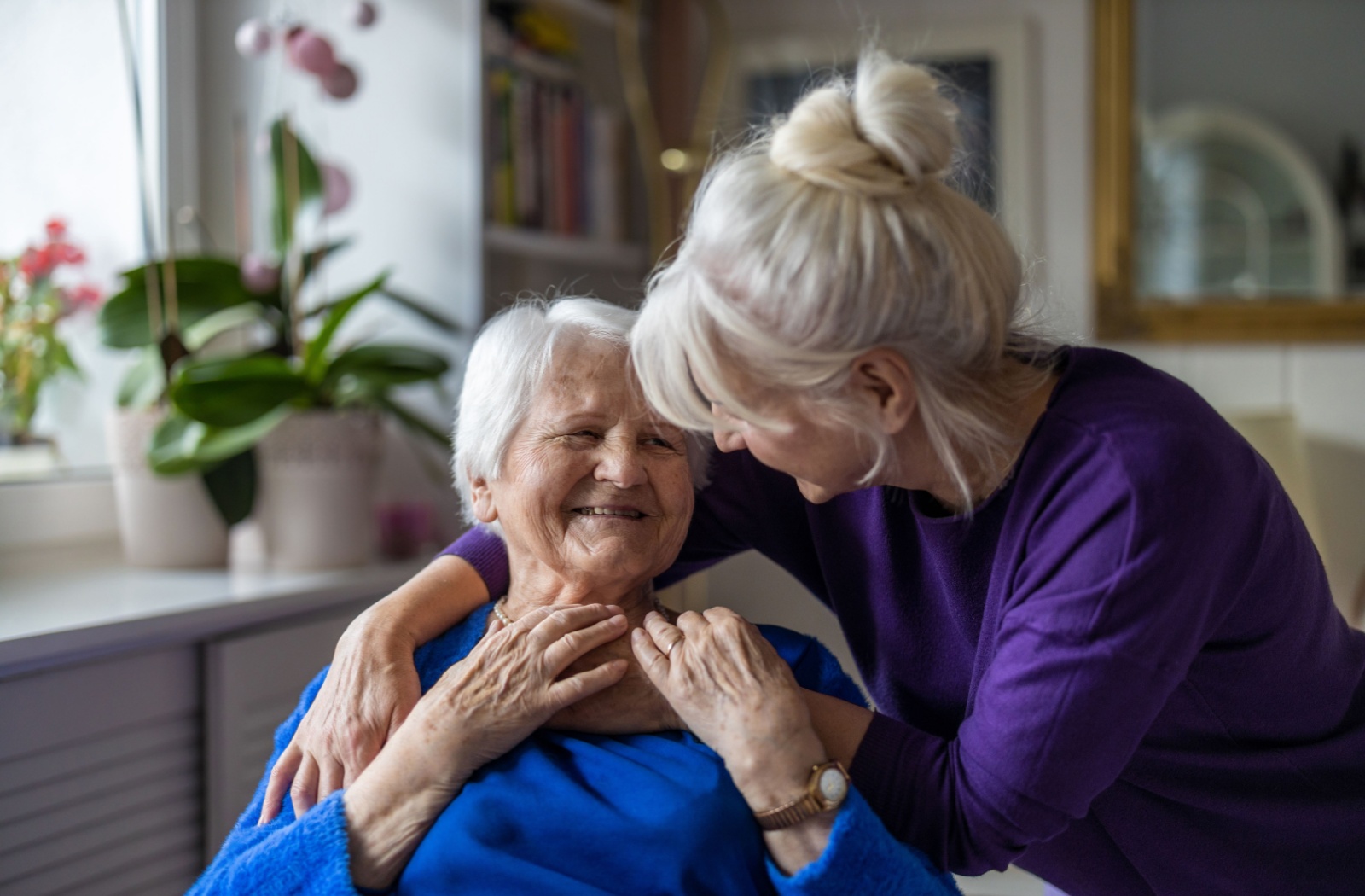 An older woman sitting down and smiling while her daughter hugs her from behind