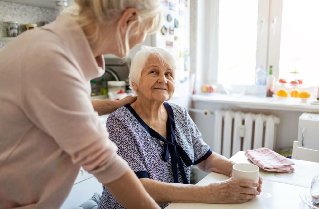 A middle-aged woman leaning on a dining table and resting her hand on the back of an older woman who is holding a coffee mug and looking up at her