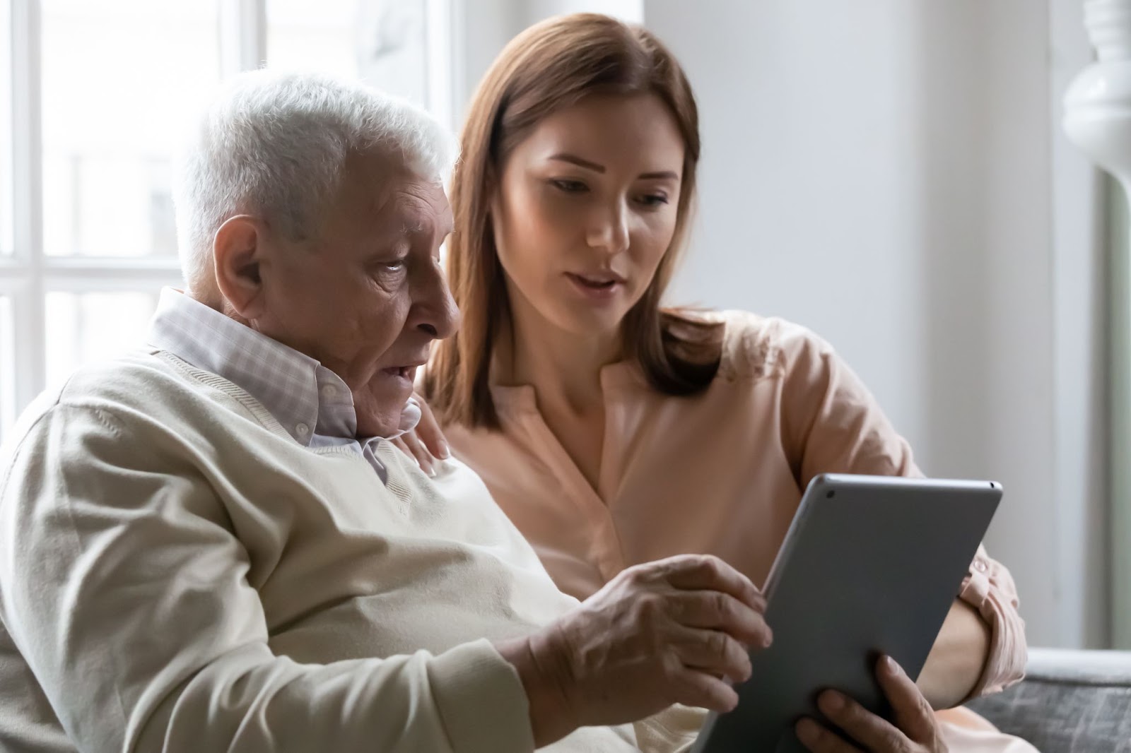 An adult daughter sitting on the couch with her older father and showing him something on a tablet as he looks on, somewhat confused