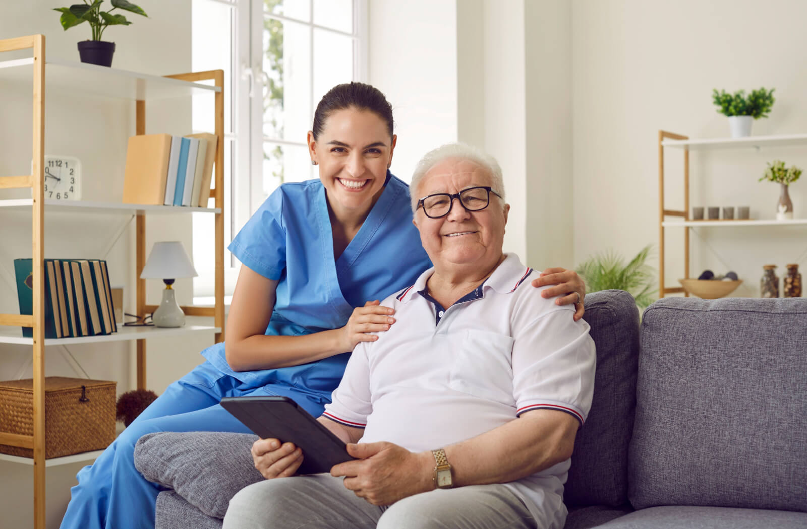 An older adult and a caregiver in assisted living, sitting on a couch and smiling at the camera while looking through a tablet.