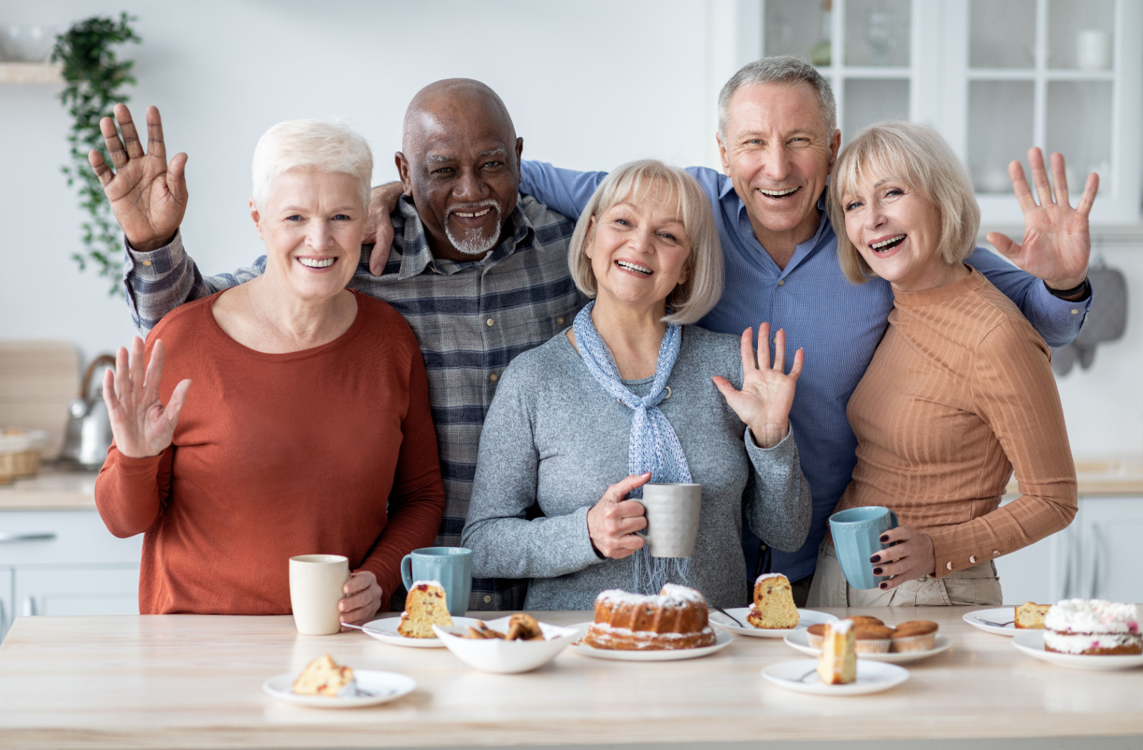 A group of older adults in assisted living smiling and waving at the camera in front of a table full of snacks.