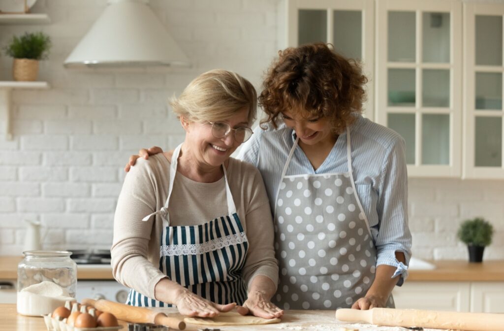 Two women wearing aprons and smiling work together at the countertop to roll out dough for a baking project.