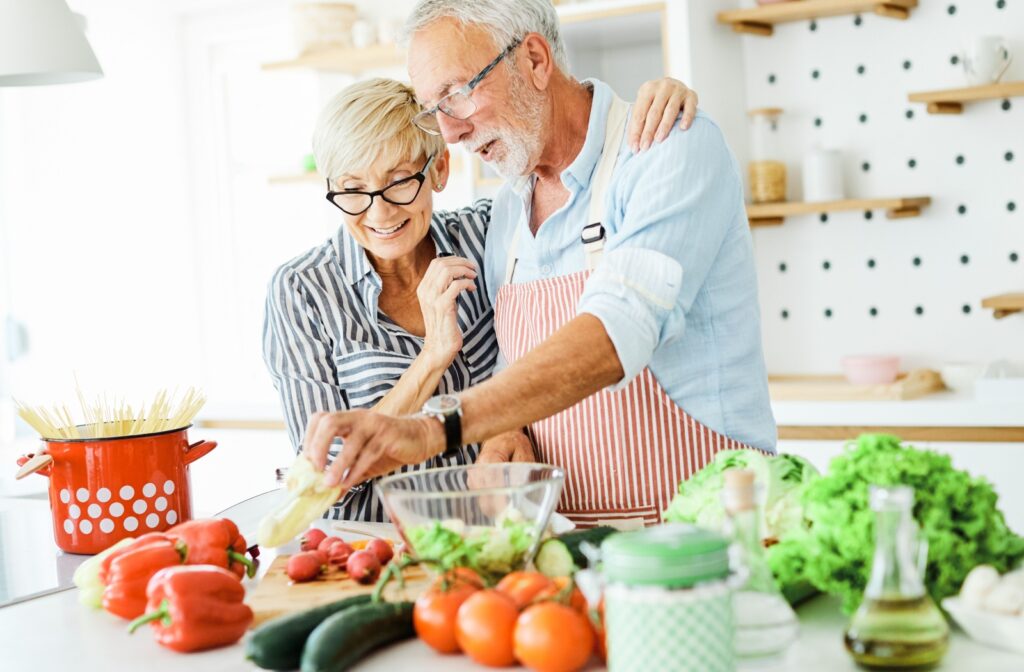 An older couple smiling while preparing assorted vegetables for a brain-friendly food in a brightly-lit kitchen.