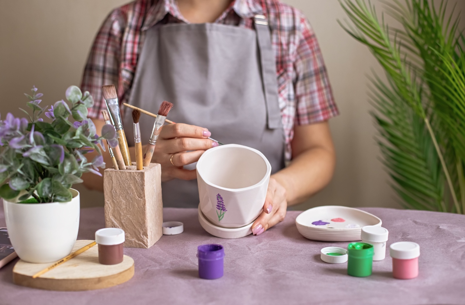 A person sitting at a craft desk, wearing an apron and painting flowers on a flower pot.