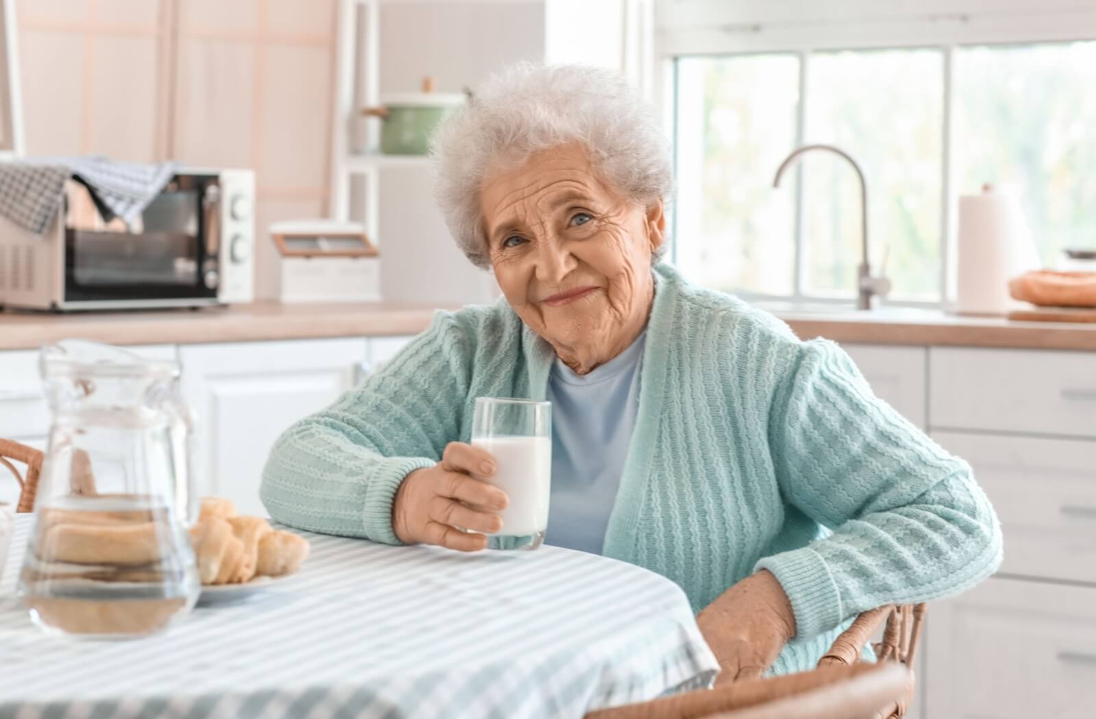 Smiling senior in a light blue sweater holding a glass of milk at a kitchen table.