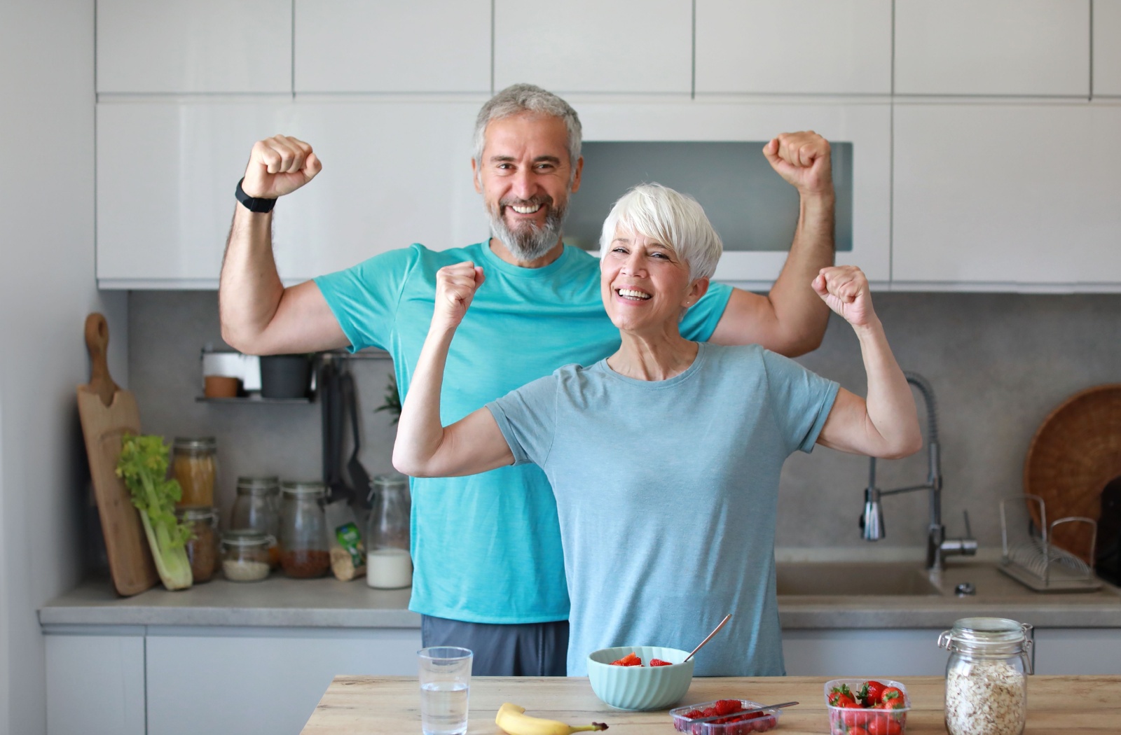 Two adults flexing their muscles demonstrating strength while eating a light fruit snack in a well lit kitchen.