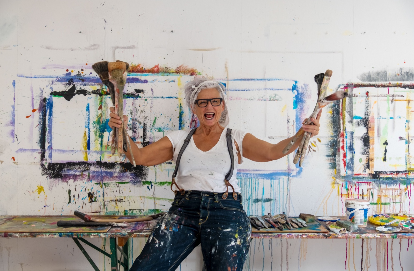 A grinning senior splattered with paint in a painting studio holds up handfuls of large paintbrushes in each hand