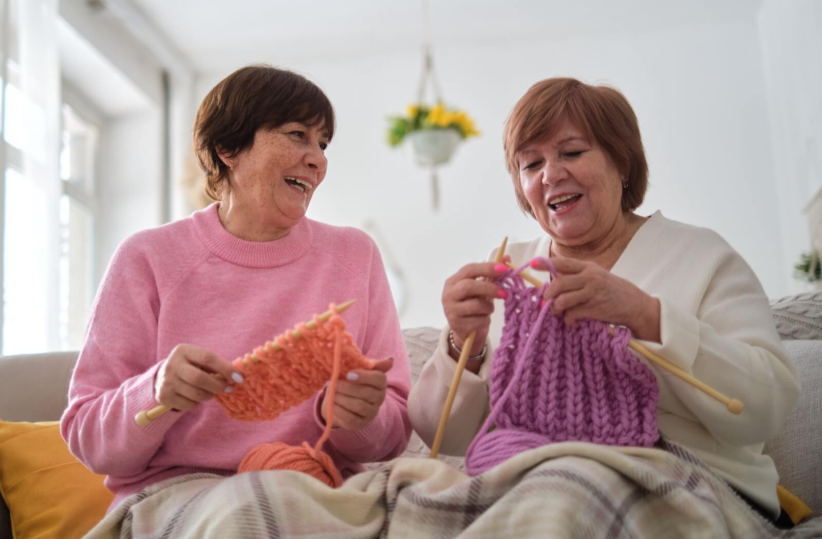 Two seniors, covered by a blanket for warmth, laugh and chat while sitting on a couch, knitting together